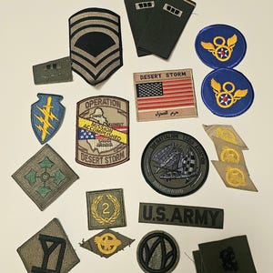 Könnte beinhalten: Eine Sammlung militärischer Abzeichen und Insignien, darunter Dienstgradstreifen, Einheitsabzeichen und das Namensband der US-Armee. Die Abzeichen weisen verschiedene Designs, Farben und Texte wie "Desert Storm" und "Operation Accomplished" auf.