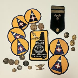 Op de afbeelding: Een verzameling militaire emblemen, knopen en insignes. Bevat ronde patches met een gele rand, een blauw-wit ontwerp en een rode ster. Bevat ook een zwarte patch met tekst en een omtrek van New Hampshire, plus diverse metalen knopen en een zwart schouderstuk.