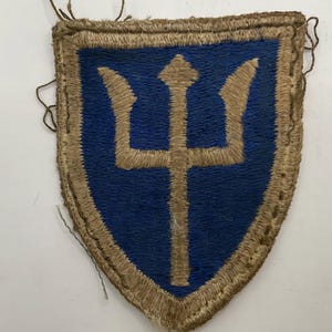 May include: A shield-shaped embroidered patch featuring a blue background with a tan trident symbol in the center. The patch has a thick tan border and visible loose threads along the edges, indicating it is a fabric military-style insignia.