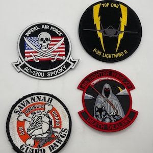 Może przedstawiać: Cztery haftowane naszywki o tematyce wojskowej. Jedna przedstawia czaszkę i flagę USA z napisem "INFIDEL AIR FORCE AC-130U SPOOKY". Inna ukazuje odrzutowiec F-35 z błyskawicami i "TOP DOG F-35 LIGHTNING II". Trzecia pokazuje buldoga z "SAVANNAH GEORGIA AIR GUARD DAWGS". Czwarta przedstawia postać w kapturze i wilka z "8th FIGHTER WING XP DEATH DEALERS".