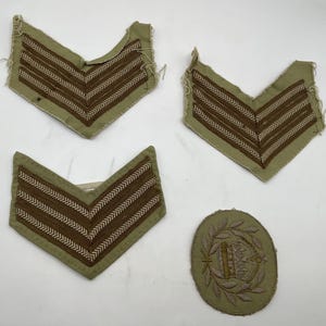 May include: Three V-shaped military rank patches in tan fabric with dark brown chevron embroidery and one oval-shaped patch featuring an embroidered crown surrounded by a laurel wreath, all displaying frayed edges and detailed stitching.