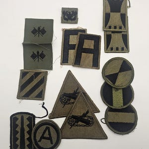 Könnte beinhalten: Eine Sammlung von olivgrünen und schwarzen gestickten Militäraufnähern in verschiedenen Formen wie Quadraten, Dreiecken, Kreisen und Rechtecken. Die Aufnäher zeigen verschiedene Symbole wie den Buchstaben A, die Buchstaben FA, eine Rakete und geometrische Muster.