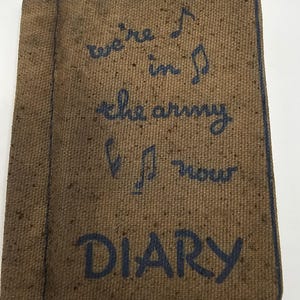 Può includere: Un diario rivestito in tessuto marrone testurizzato con testo ricamato in blu scuro che recita "we're in the army now" accompagnato da simboli di note musicali, e la parola "DIARY" cucita in grandi lettere blu scuro in basso.