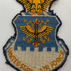 Può includere: Una toppa ricamata della United States Air Force con un'aquila dorata in alto, uno scudo blu con un emblema alato dorato centrale e una base bianca con il testo UNITED STATES AIR FORCE ricamato in filo dorato lungo il bordo inferiore.