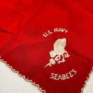 以下が含まれることがあります： U.S. NAVYおよびSEABEESという白い刺繍文字と、工具を持った蜂の白い刺繍エンブレムが中央に施された鮮やかな赤色の布製スクエア。縁は装飾的な白いスカラップステッチで仕上げられています。