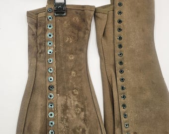 May include: A pair of olive drab canvas gaiters featuring a vertical row of metal eyelets along the front edge, a top buckle closure, and a fabric strap. The material shows signs of wear, discolouration, and oxidation on the metal hardware.