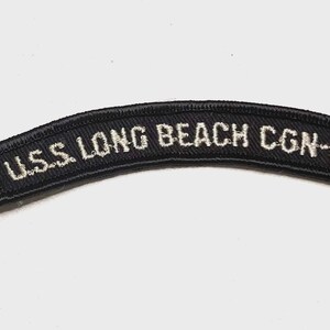 May include: A curved, black embroidered patch featuring white stitched text that reads U.S.S. LONG BEACH CGN-9. The patch has a thick, black stitched border and a textured fabric surface, designed for attachment to clothing or gear.