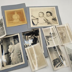 May include: A collection of vintage black and white and sepia-toned photographs mounted on blue cardstock. The images depict various scenes including portraits of infants, a person holding a baby, outdoor landscapes, and architectural structures.