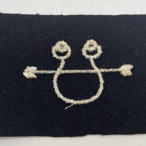 May include: A rectangular black felt patch featuring an embroidered silver-colored design. The embroidery depicts a horizontal arrow intersecting a circular shape that loops upward into two small, rounded, knot-like forms at the top center.
