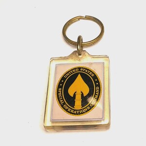 May include: A rectangular clear acrylic keychain featuring a circular emblem of the United States Special Operations Command. The emblem displays a yellow spearhead on a black background, surrounded by a gold border with black text.