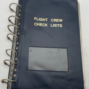 Könnte beinhalten: Ein dunkelblauer Ringbuchordner mit dem in Gold aufgedruckten Text FLIGHT CREW CHECK LISTS auf der Vorderseite. Der Ordner verfügt über eine durchsichtige rechteckige Kunststoffhülle im unteren Bereich und silberne Metallringe an der linken Seite.
