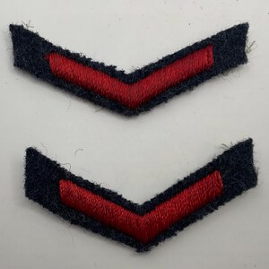 May include: Two chevron-shaped military rank patches featuring a dark navy blue felt base with a vibrant red embroidered stripe running through the center. These V-shaped insignia pieces are identical in design, showcasing a textured fabric finish.