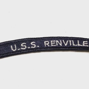 May include: A curved, dark navy blue embroidered patch featuring the white text U.S.S. RENVILLE. The patch has a thick, stitched border and a rectangular shape with slightly rounded ends, designed for attachment to a garment or uniform.