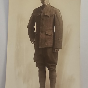 May include: Sepia-toned photograph of a man in a military uniform. He wears a dark brown jacket with pockets, matching breeches, and tall socks. A peaked cap sits atop his head. The image is a full-length portrait.