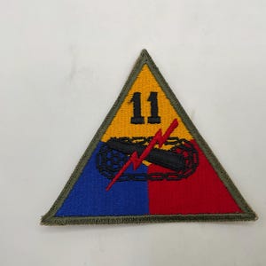 May include: A triangular embroidered patch featuring a yellow top section with the black number 11, a blue bottom left section, and a red bottom right section. A black tank tread graphic with a red lightning bolt overlay sits in the center.