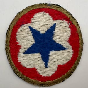 May include: A circular embroidered patch featuring a central blue five-pointed star set against a white flower-like shape, all enclosed within a red circular field and finished with a thick, gold-coloured stitched border around the outer edge.