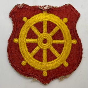 May include: A shield-shaped embroidered patch featuring a bright yellow ship's wheel centred on a solid red background. The patch has a thick red stitched border and shows visible fraying along the edges of the fabric backing.
