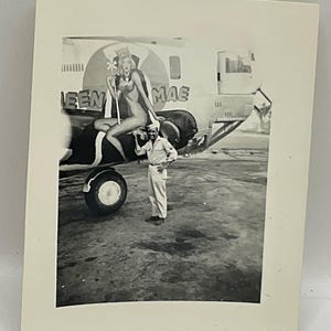 May include: A black and white photograph showing a person in a military uniform standing next to an airplane. The aircraft features nose art of a woman wearing a crown and holding a scepter, with the text QUEEN MAE painted on the side of the fuselage.