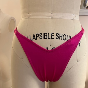Puede incluir: Un bañador de color rosa brillante con una pierna de corte alto y un frente en forma de V. El bañador está en un maniquí blanco.