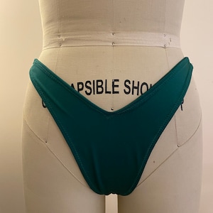 Puede incluir: Un bikini de color verde azulado con la parte delantera en forma de V y una pierna de corte alto. La parte inferior del bikini está en un maniquí blanco.