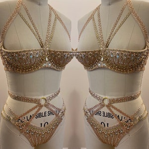 Puede incluir: Un conjunto de lencería de color dorado con un diseño delicado e intrincado. El sujetador y la braga están adornados con brillantes pedrería y presentan un diseño de tirantes cruzados.