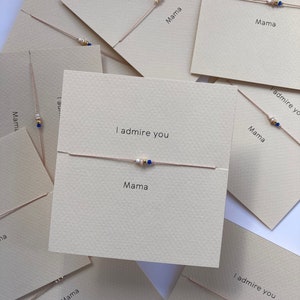 May include: Several beige cards with the phrases "I admire you" and "Mama" printed in black. Each card displays a delicate bracelet with gold, white, and blue beads on a thin, light-coloured cord. The bracelets are the main focus of the image.