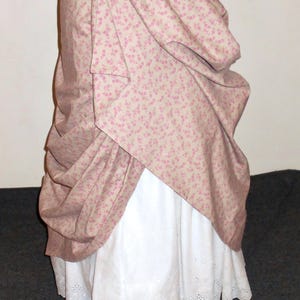 May include: A historical-style skirt on a mannequin. The skirt features a light pink floral patterned overskirt draped over a white eyelet underskirt. The fabric is gathered and draped, showcasing the design.