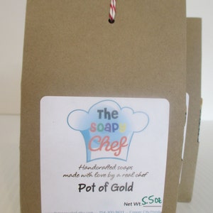 May include: Brown paper bag with a white label featuring a chef hat logo and the text "The Soap Chef." The label reads "Pot of Gold" and "Handcrafted soaps made with love by a real chef." The net weight is 5.5 oz. A red and white string is attached.