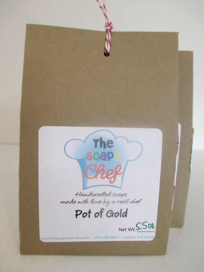 May include: Brown paper bag with a white label featuring a chef hat logo and the text "The Soap Chef." The label reads "Pot of Gold" and "Handcrafted soaps made with love by a real chef." The net weight is 5.5 oz. A red and white string is attached.