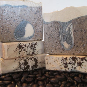 May include: Four bars of handmade coffee soap stacked on top of each other. The soap is a light brown color with dark brown swirls and specks of coffee grounds. The soap is sitting on a bed of coffee beans.