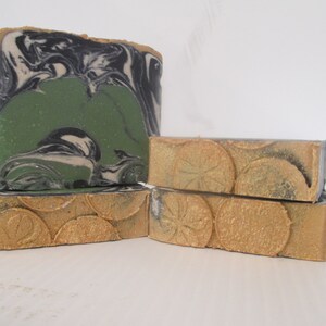 May include: Artisan soap bars in various sizes. One large bar features a green center with black and white swirls. Smaller bars have a gold-colored, textured design. The soaps are stacked, showcasing their unique textures and colors.