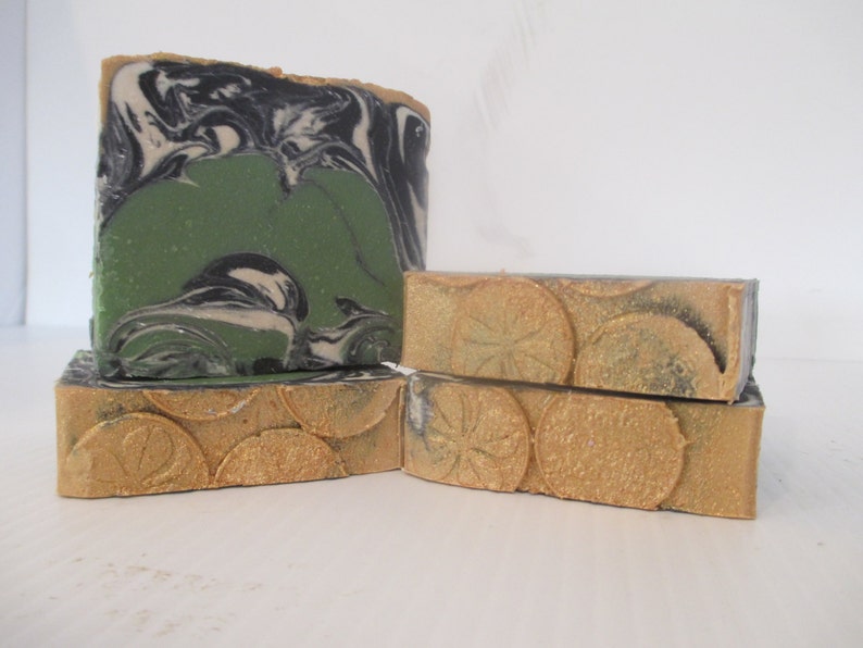 May include: Artisan soap bars in various sizes. One large bar features a green center with black and white swirls. Smaller bars have a gold-colored, textured design. The soaps are stacked, showcasing their unique textures and colors.