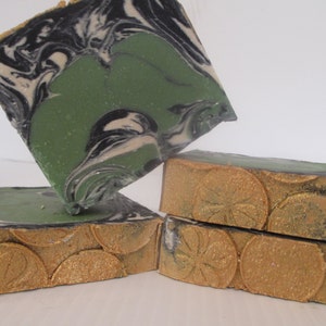 May include: Four bars of handmade soap. The top of the soap has a green, black, and white swirl design. The sides are a gold color with a circular pattern. The soap is stacked on top of each other.