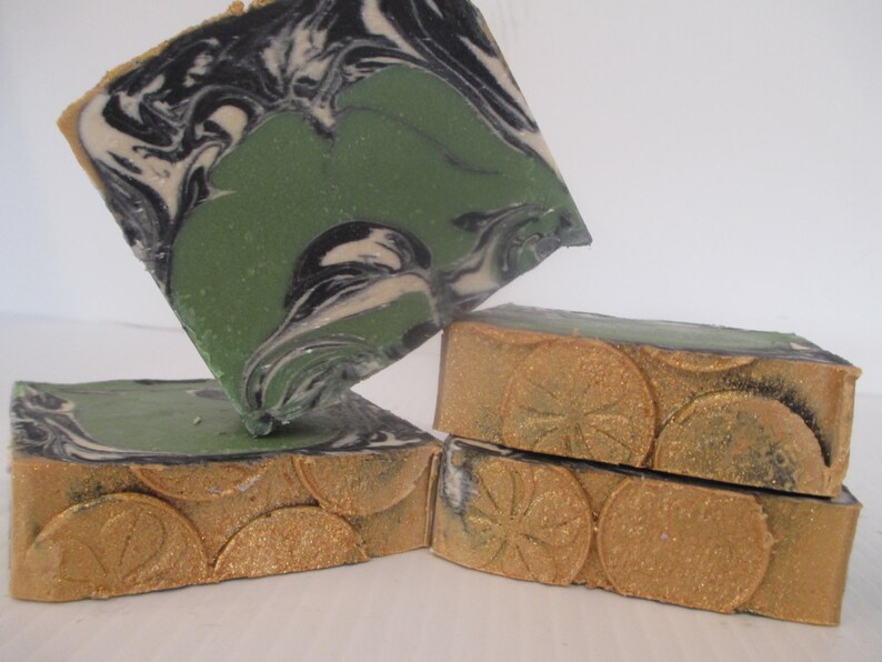 May include: Four bars of handmade soap. The top of the soap has a green, black, and white swirl design. The sides are a gold color with a circular pattern. The soap is stacked on top of each other.