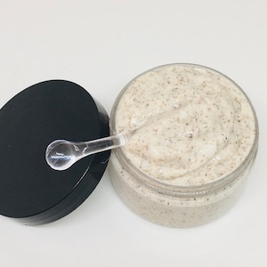 May include: A clear jar with a black lid filled with a white and brown exfoliating scrub. A clear plastic spoon is resting on the top of the scrub.