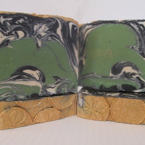 May include: Handmade soap bars with a unique design. The top of the soap features a green center with black and white swirls. The bottom of the soap has a gold-colored, textured design. The soap is cut into rectangular bars.