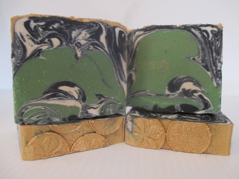 May include: Handmade soap bars with a unique design. The top of the soap features a green center with black and white swirls. The bottom of the soap has a gold-colored, textured design. The soap is cut into rectangular bars.