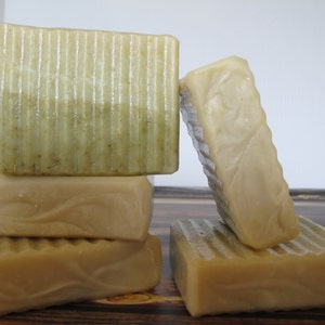 May include: A stack of four handmade soap bars. The top bar is green and white with a layered design. The other three bars are a solid beige color. The bars are arranged on a wooden surface.