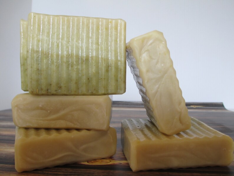 May include: A stack of four handmade soap bars. The top bar is green and white with a layered design. The other three bars are a solid beige color. The bars are arranged on a wooden surface.