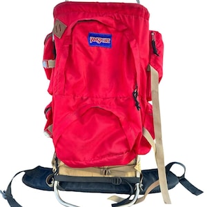 May include: A bright red JanSport external frame backpack featuring a metal frame, a blue brand patch, side pockets, and tan adjustable straps. The pack includes a black padded waist belt and black zipper pulls, with a distinct rectangular shape.