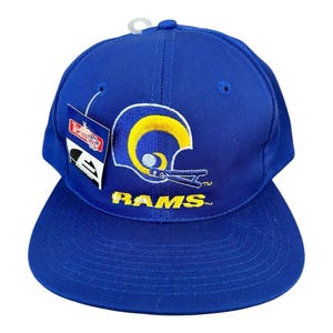 May include: Blue and yellow baseball cap with a Rams logo and the word "RAMS" embroidered on the front. The cap has a Team Issue tag attached to the side.