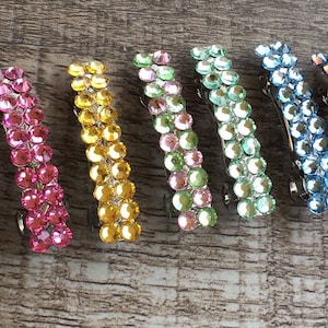 May include: A set of 12 hair clips with a variety of colors, including clear, pink, yellow, green, blue, and purple. Each clip is adorned with rows of sparkling rhinestones.