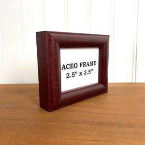 May include: A dark brown wooden ACEO frame, measuring 2.5 x 3.5 inches, is displayed on a wooden surface. The frame has a rectangular shape with a slightly curved edge. The text "ACEO FRAME 2.5" x 3.5"" is printed on the white insert.