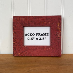 May include: A red, textured ACEO frame with the text "ACEO FRAME 2.5" x 3.5"" in the center. The frame is square and has a distressed finish, with visible wood grain and a white interior.