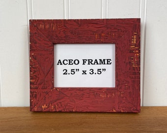 May include: A red, textured ACEO frame with the text "ACEO FRAME 2.5" x 3.5"" in the center. The frame is square and has a distressed finish, with visible wood grain and a white interior.