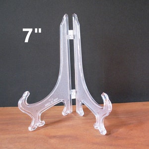 May include: A clear plastic display stand, 7 inches tall, with a curved, triangular base and two prongs for holding objects.