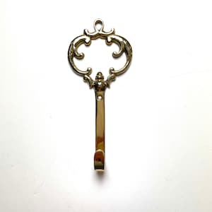 May include: A gold-toned key-shaped hook with a decorative, ornate design. The hook is made of metal and has a smooth, polished finish.