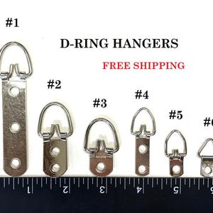 May include: Six different sizes of silver D-ring hangers for hanging pictures or other items. The hangers are numbered 1 through 6 and are shown against a ruler.