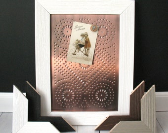 May include: A white framed copper-colored metal wall decor with a vintage postcard and decorative button. The copper panel has a punched design. Several frame corner samples in various colors are in front of the frame.
