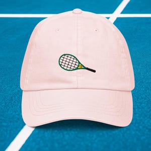 May include: A pink baseball cap with an embroidered green tennis racket and yellow tennis ball.